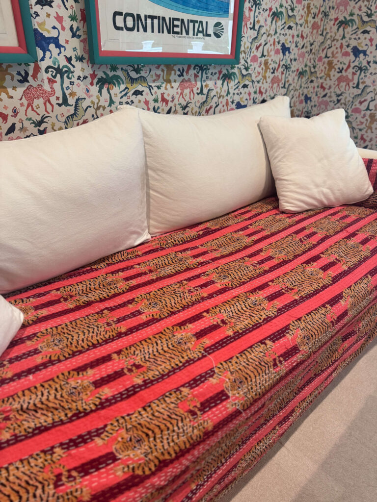 A vibrant Indian textile with tiger motifs in red, orange, and black tones covers a sofa with white cushions, set against colorful animal-patterned wallpaper.