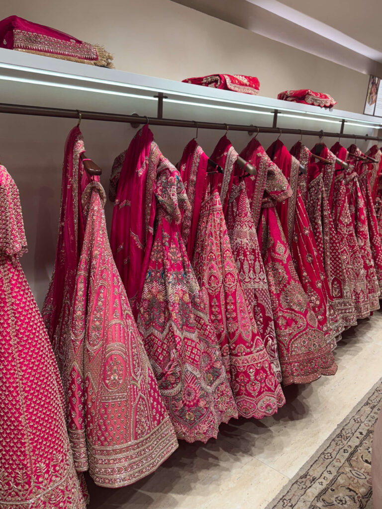 A row of intricately embroidered red and pink bridal lehengas hanging on a rail in a boutique, showcasing traditional Indian craftsmanship.