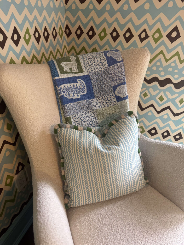 Cozy corner with a white boucle armchair, patterned cushion, and blue and green animal-print quilt from Dastkar Ranthambore, India against bold geometric wallpaper.
