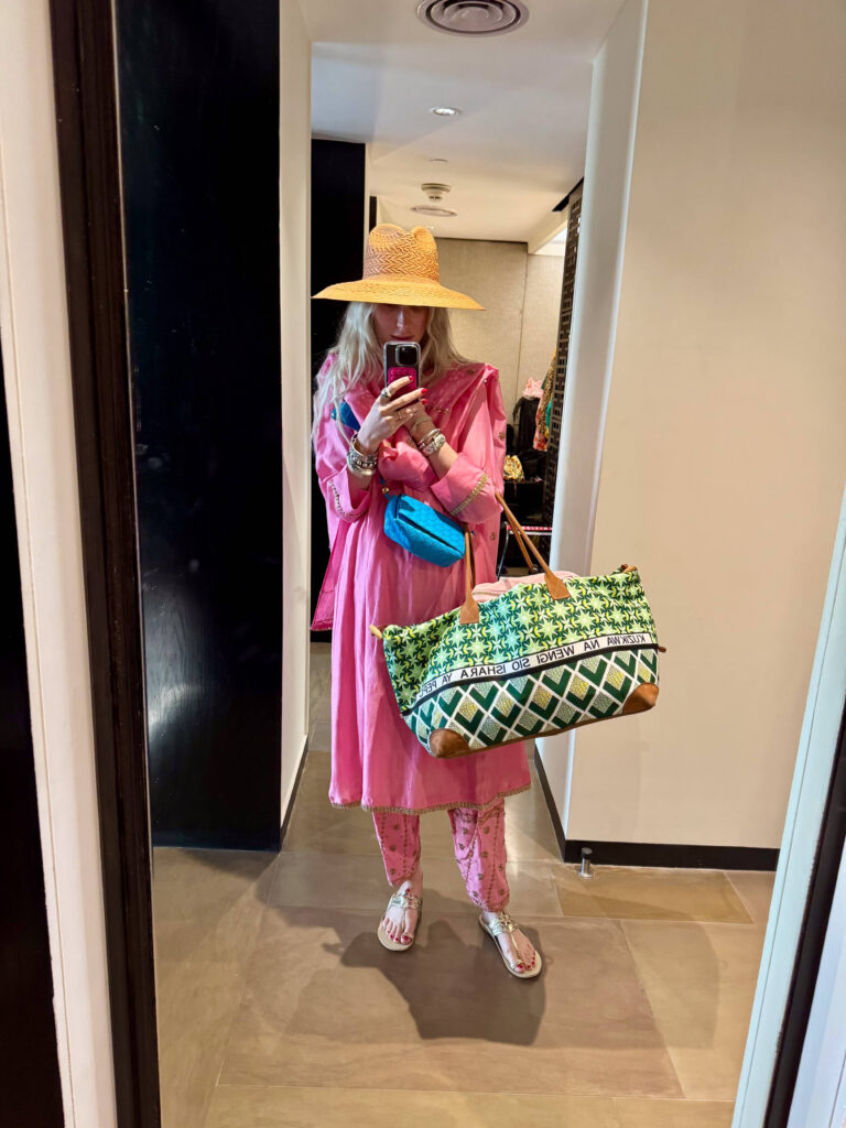 Woman in pink Indian outfit with straw hat taking mirror selfie in hotel hallway, holding green patterned weekender bag and wearing gold sandals.