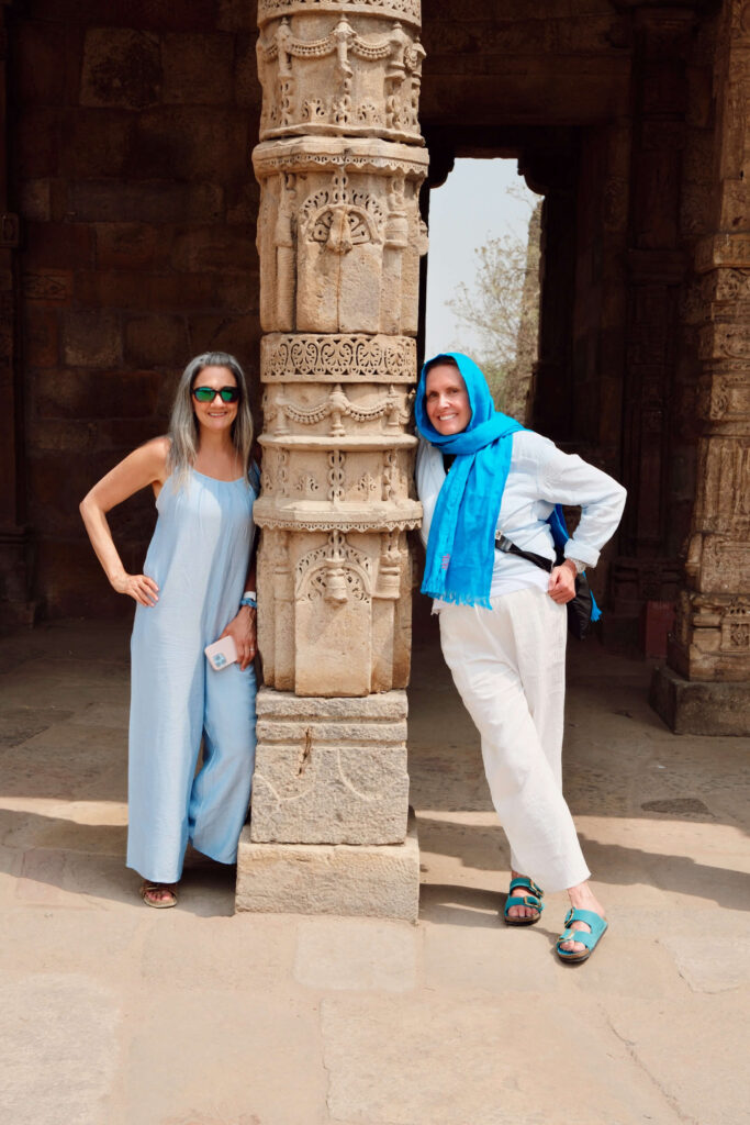 Two women posing on either side of an intricately carved stone column at a historic site, one in a light blue jumpsuit, the other in white outfit with bright blue scarf.