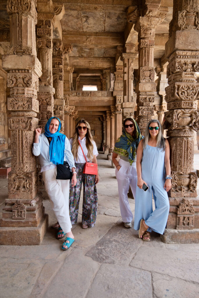 Four women standing among carved stone pillars in a historic Indian site (Quatab Minar), wearing colorful and airy travel outfits, two with scarves covering their heads.