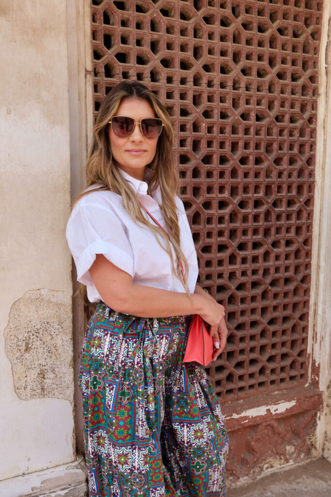 Woman wearing sunglasses, white shirt, and printed wide-leg pants poses in front of traditional Indian jali (lattice) window.