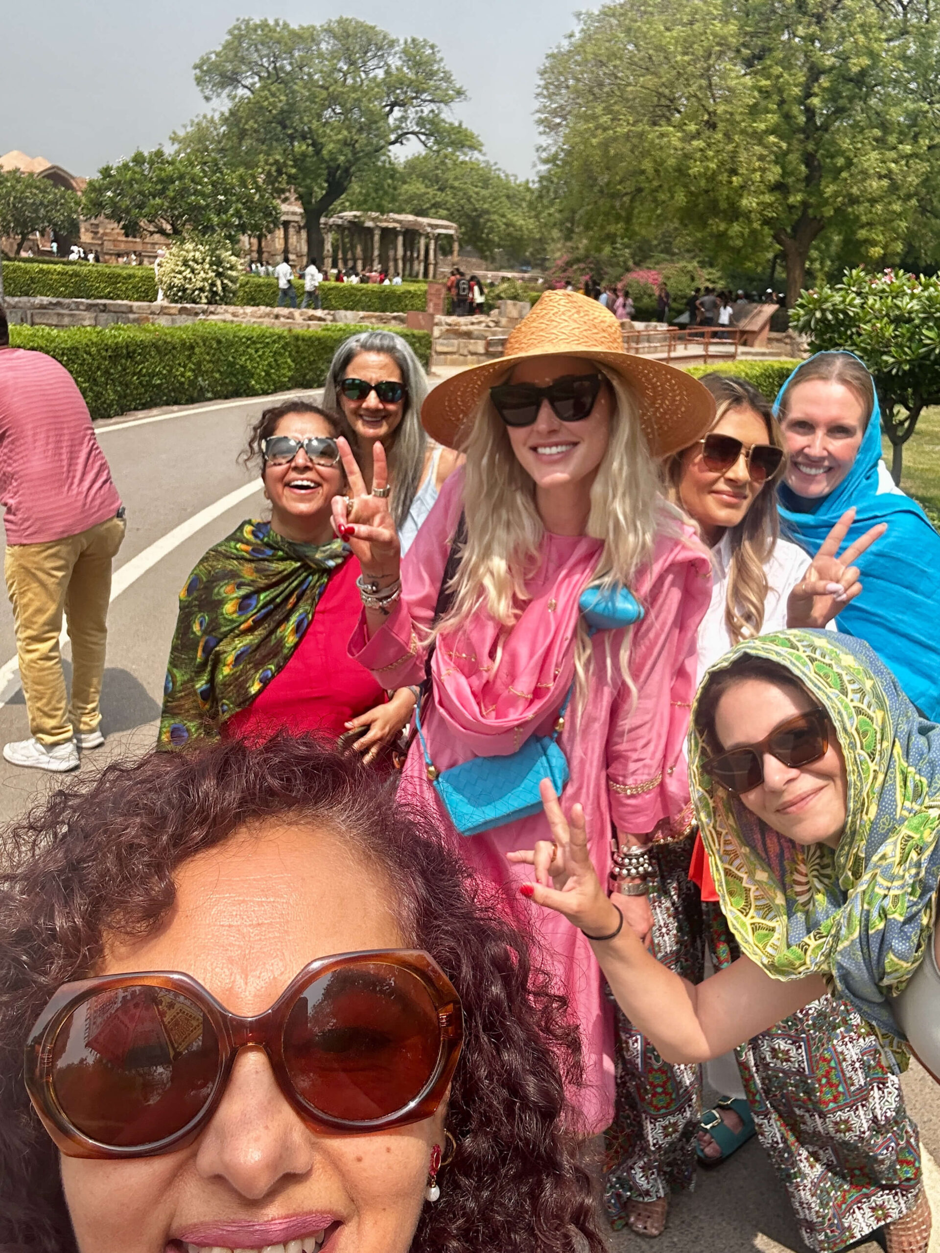 Group of seven smiling women posing outdoors in India, wearing colorful scarves, sunglasses, and modest outfits on a sightseeing tour.