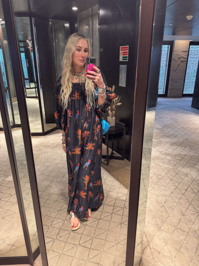 Woman taking mirror selfie in a hotel hallway, wearing a black maxi dress with vibrant palm tree and parrot print, accessorized with silver sandals and statement Chanel jewelry.