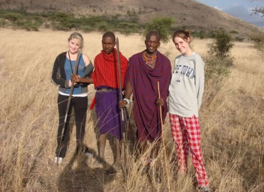Two young women and two Maasai men standing together in tall grass in rural Kenya, smiling and holding spears