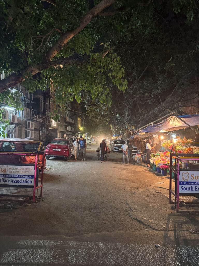 A bustling street scene at night in India, with vendors selling colorful flowers under a canopy and people walking down a narrow lane lined with shops and trees.