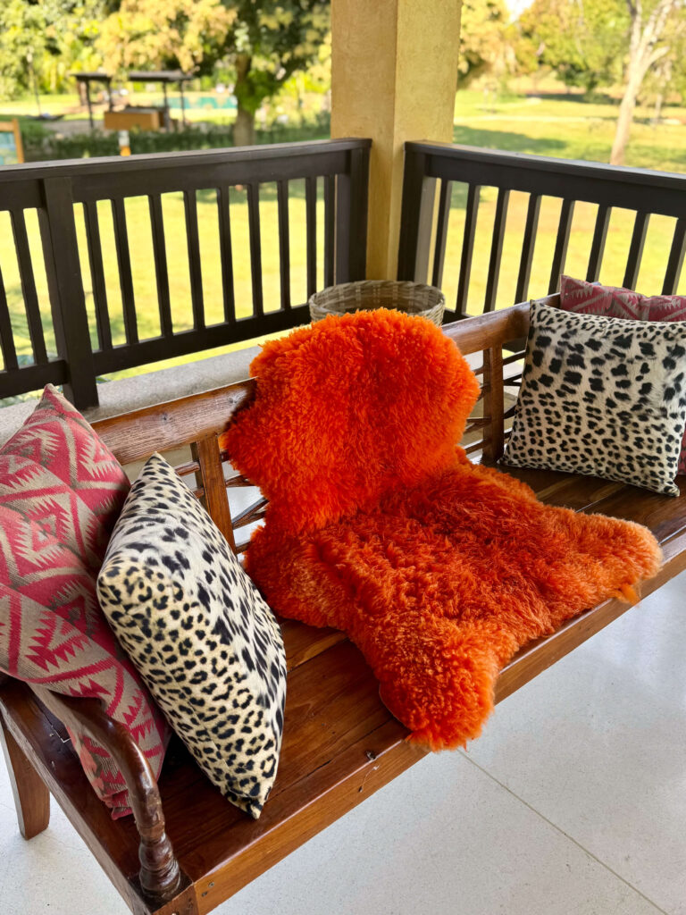 Wooden porch bench decorated with bold, patterned cushions and a bright orange sheepskin, overlooking a garden.