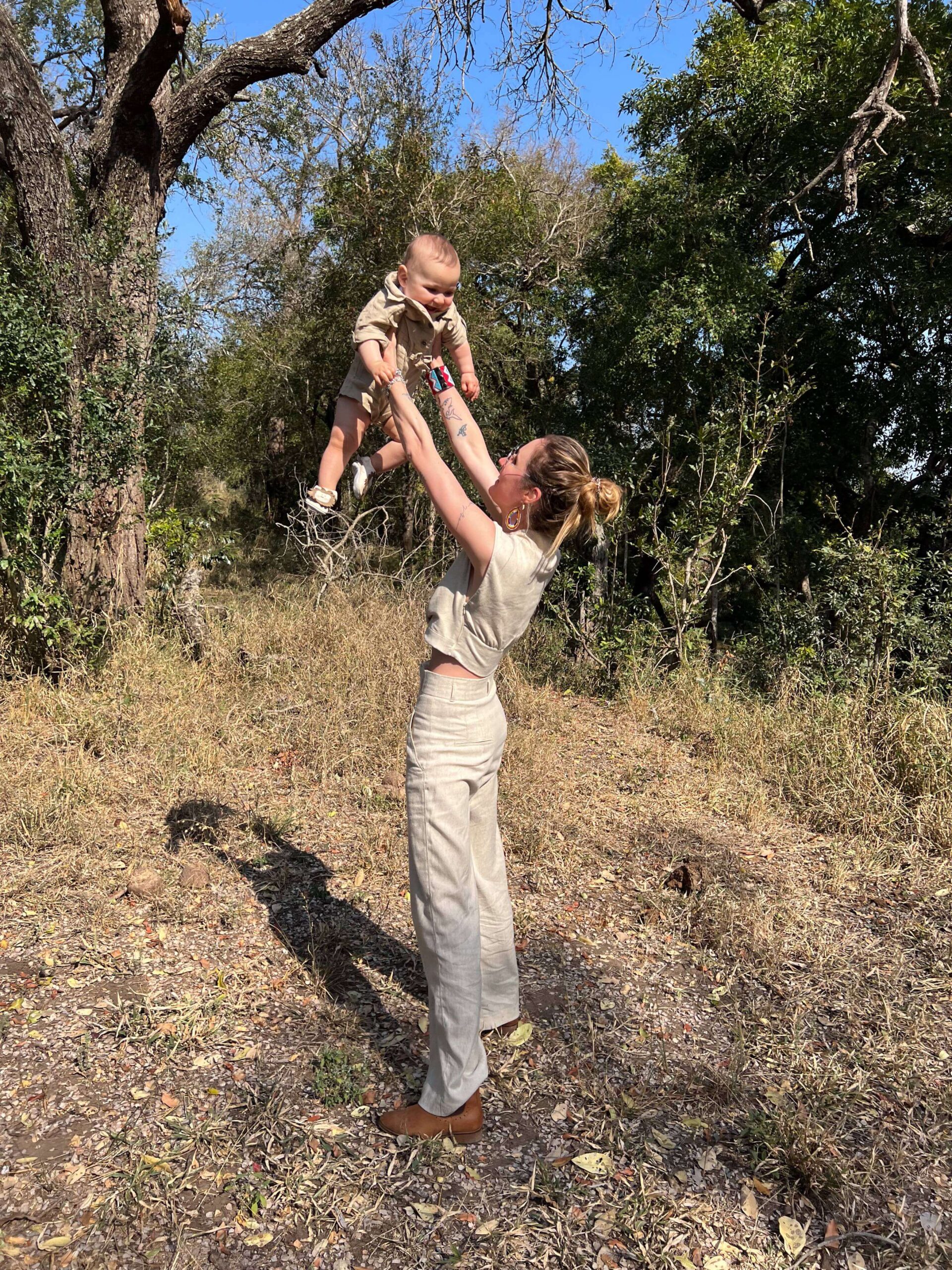 Mother lifting her baby joyfully into the air during a safari walk in the African bush, both dressed in neutral safari outfits
