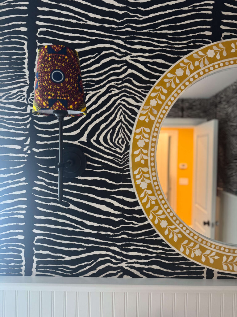 A bold interior wall featuring black-and-white zebra print wallpaper, a circular yellow mirror with a white floral pattern, and a colorful African fabric lampshade.