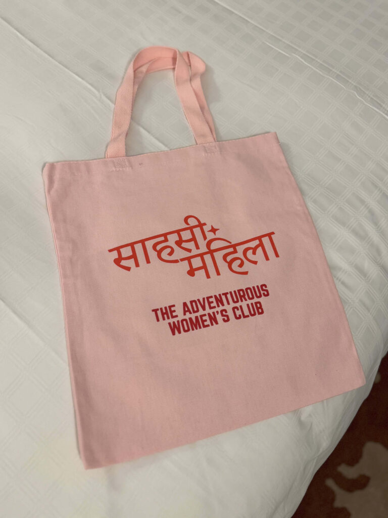 A light pink canvas tote bag with bold red Hindi and English text reading “The Adventurous Women’s Club,” laid out on a white bedspread.