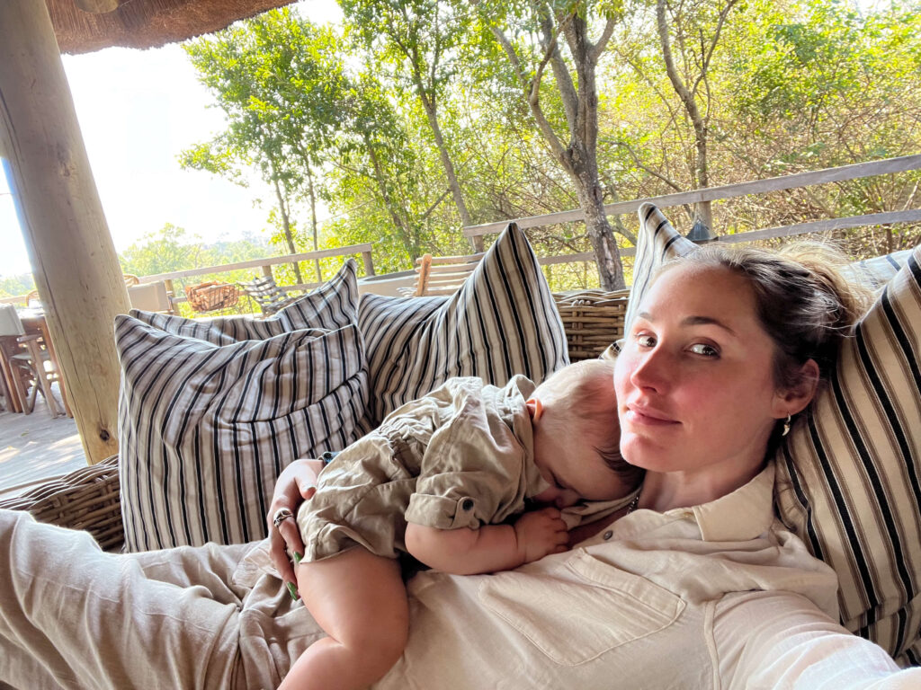 Mother reclining on an outdoor couch with her baby sleeping on her chest, surrounded by striped cushions and nature in the background