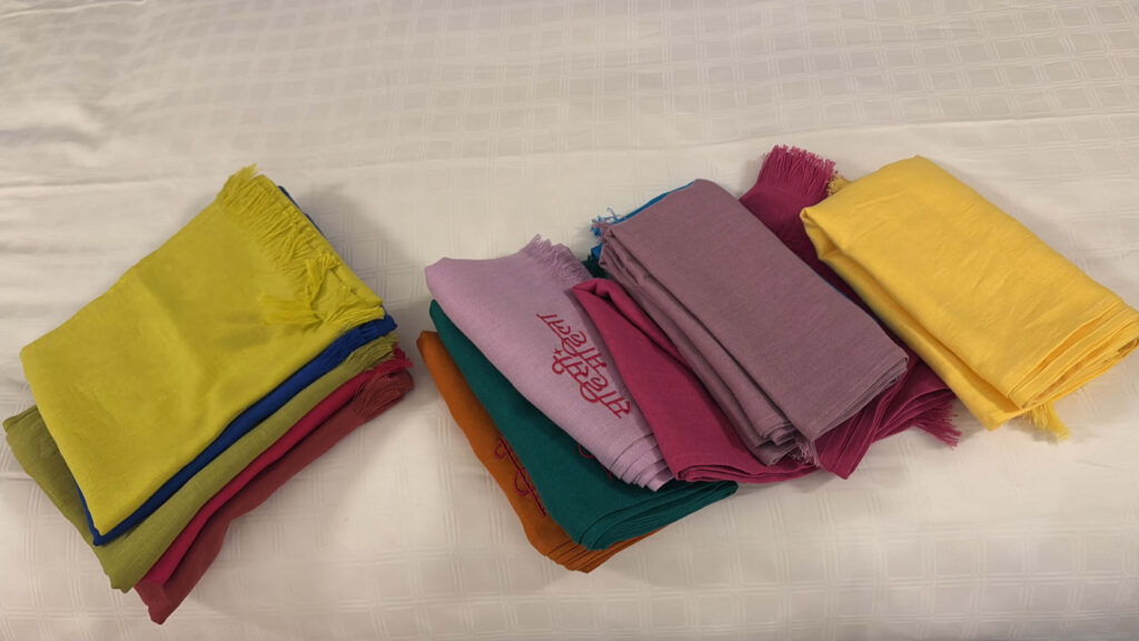 A collection of folded scarves in vibrant colors like mustard, pink, green, and purple, some featuring embroidered Hindi lettering, neatly arranged on a bed.
