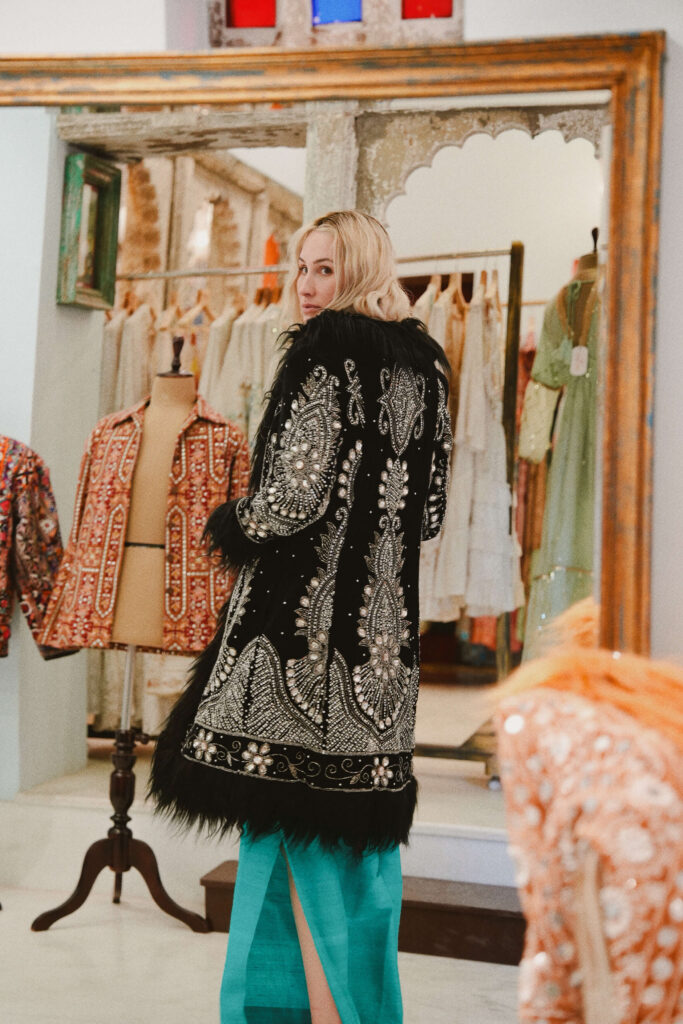 A woman wearing an intricately embroidered black velvet jacket with feather trim stands in a boutique surrounded by traditional Indian garments.