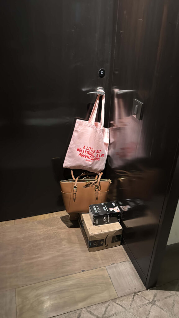 A pink tote bag hanging from a dark-colored door handle with the phrase “A little bit Bollywood, a lot adventurous” printed in red, next to a tan handbag and packages on the floor.