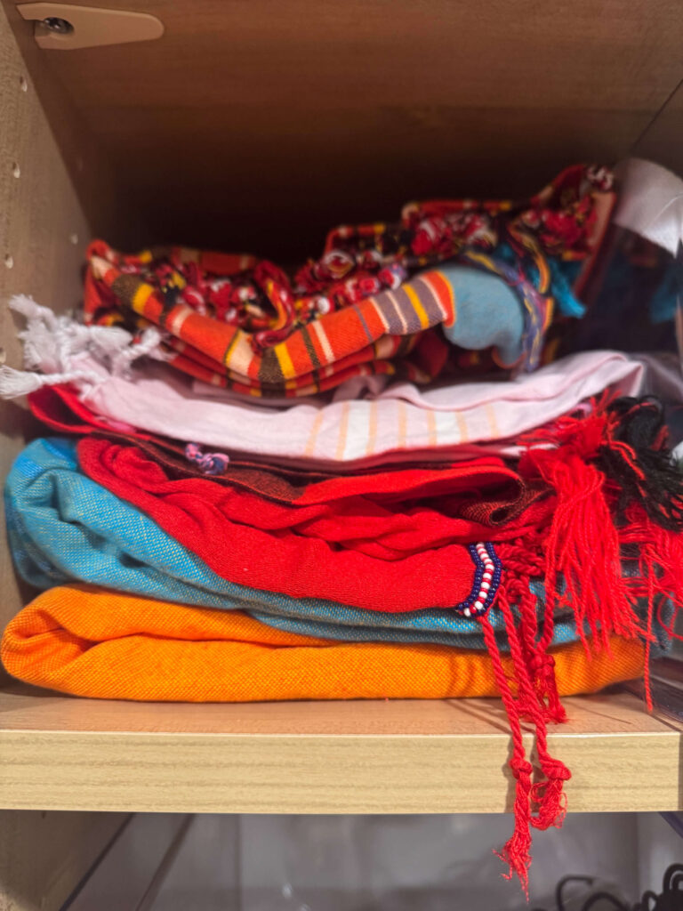A neatly stacked collection of colorful kikoi scarves from Kenya and vibrant textiles from India on a wooden shelf.