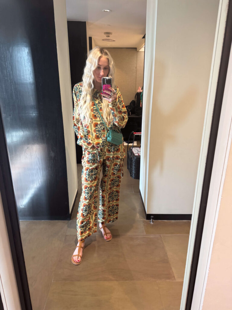 A woman taking a mirror selfie in a hotel room, wearing a colorful printed matching shirt and pants set (from Coniglio and Vivrelle) with long blonde hair, holding a pink phone.