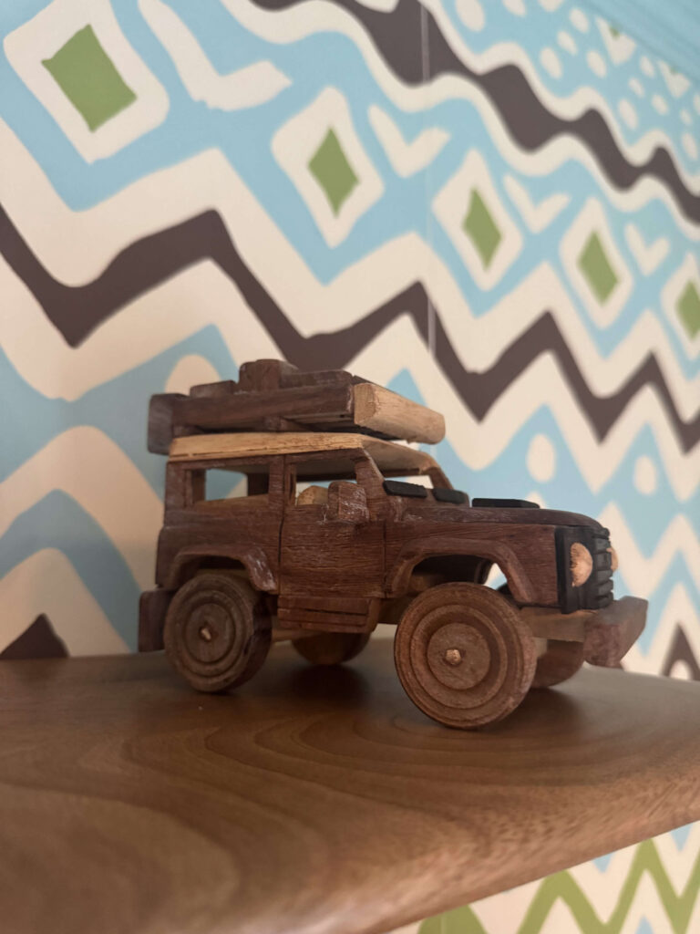 A handmade wooden toy safari truck displayed on a wooden shelf with bold blue, green, and black patterned wallpaper in the background.