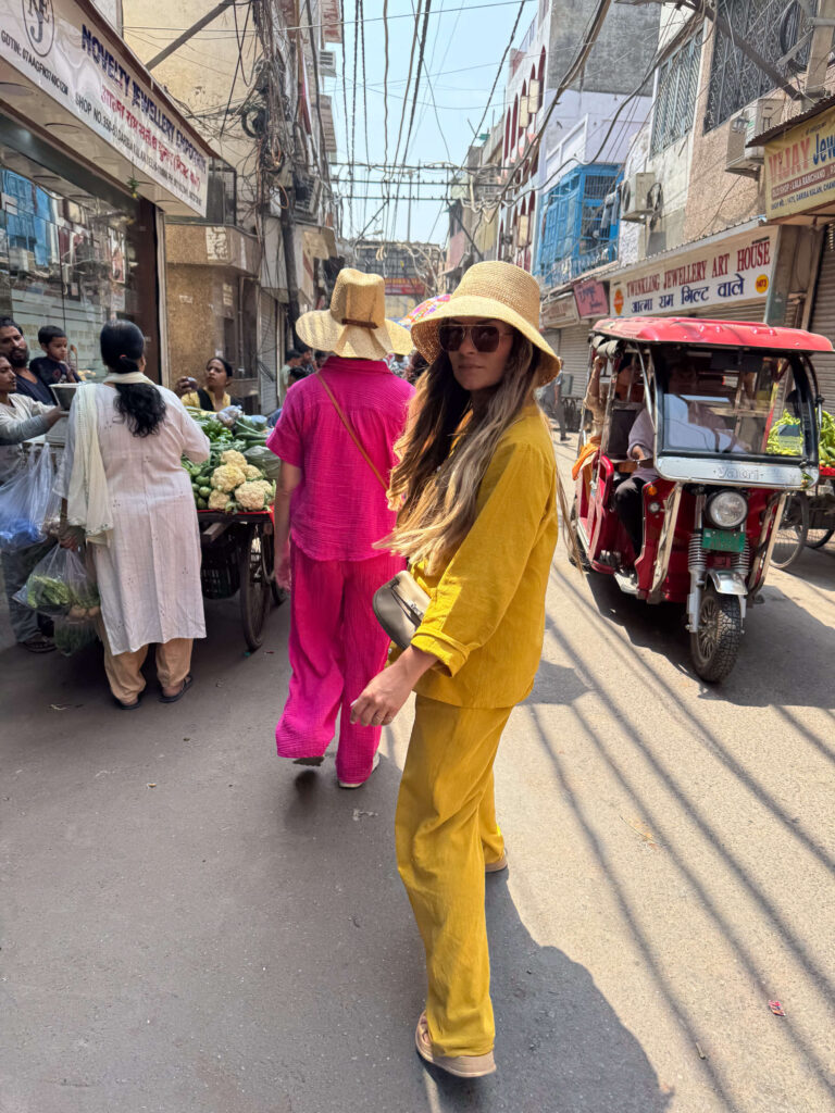 Two women dressed in bright linen sets walking through a crowded Old Delhi street with vendors, rickshaws, and hanging wires overhead.