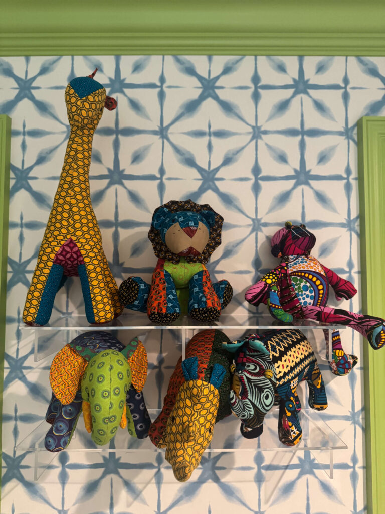 A collection of colorful handmade African animal plush toys, including a giraffe, elephant, rhino, and lion, displayed on wall-mounted acrylic shelves.