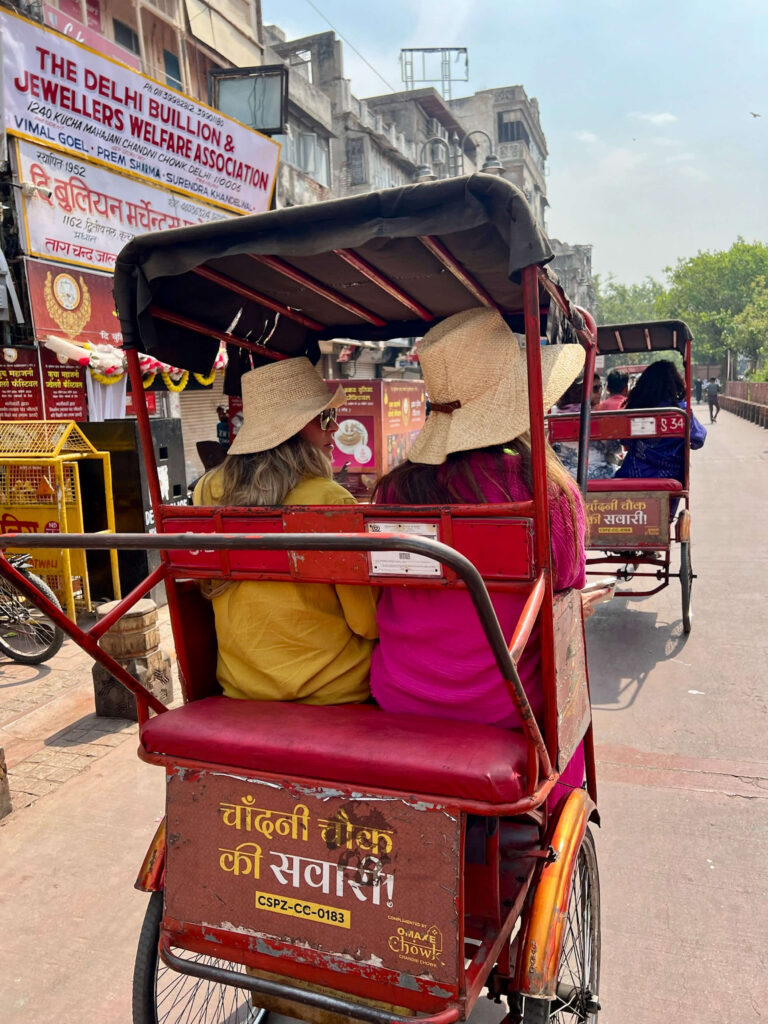 Two women in colorful outfits and wide-brimmed hats seated on the back of a red bicycle rickshaw, riding through the streets of Delhi.