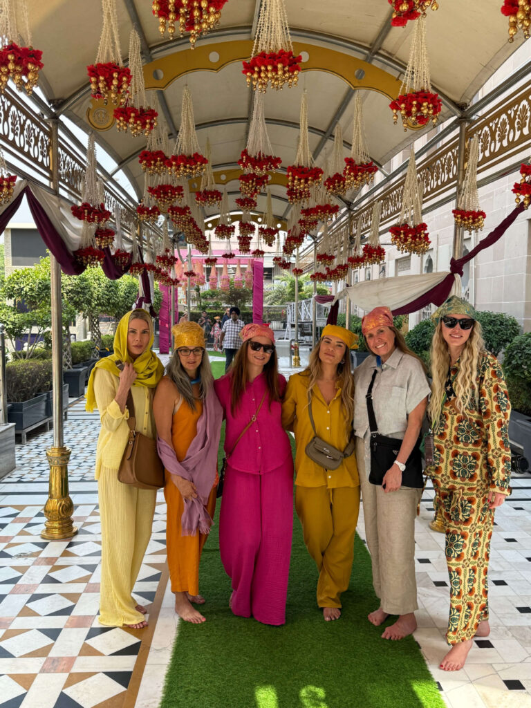 A group of six women standing barefoot under an ornately decorated canopy in India, dressed in coordinated colorful outfits and turbans, smiling for the camera.